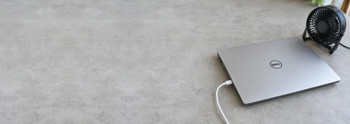 A closed Dell laptop charging beside a small desk fan on a concrete-effect workbench