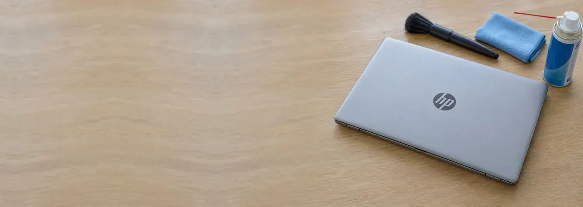 A closed HP laptop beside a brush, a cloth and a can of compressed air on an oak desk top