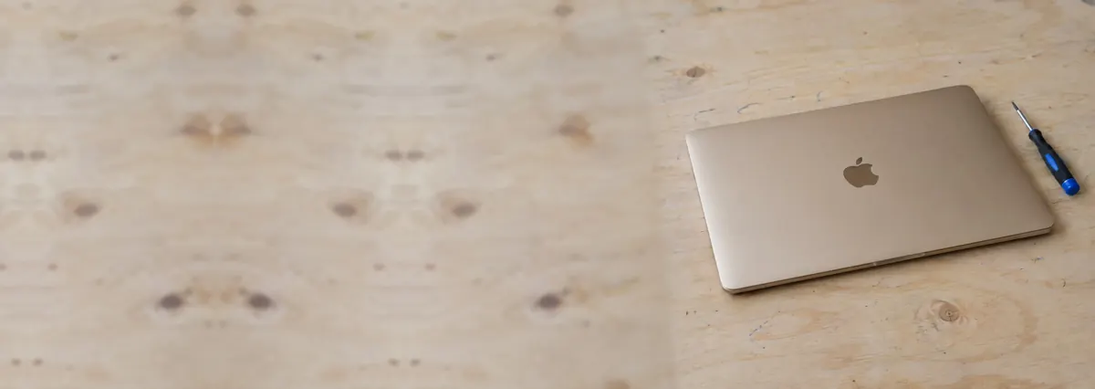 A closed champagne-gold Apple MacBook beside a screwdriver on a plywood bench