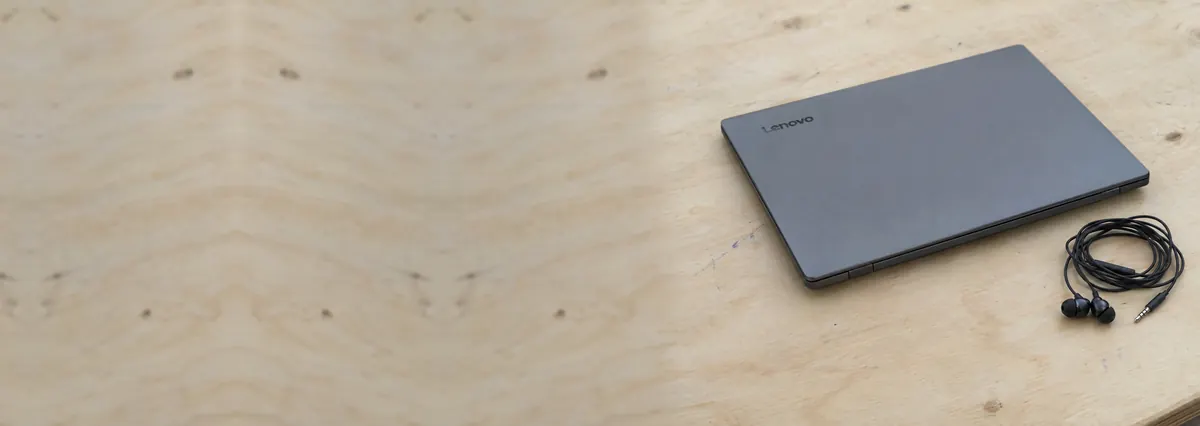 A closed grey Lenovo laptop beside coiled earphones on a plywood bench