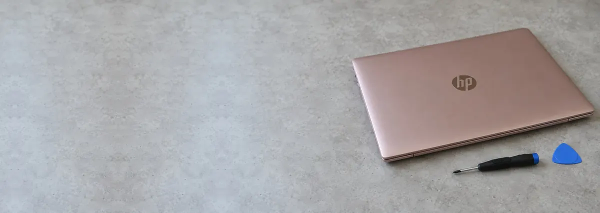 A closed rose-gold HP laptop on a concrete-effect workbench beside a screwdriver and an opening pick
