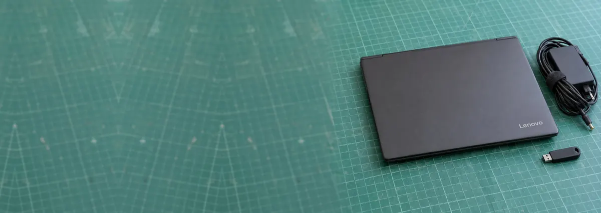 A closed graphite Lenovo laptop beside a charger cable and a flash drive on a green cutting mat