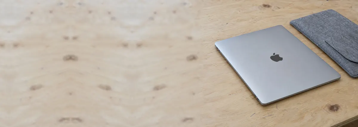 A thin silver Apple MacBook lying closed beside a grey felt sleeve on a plywood bench