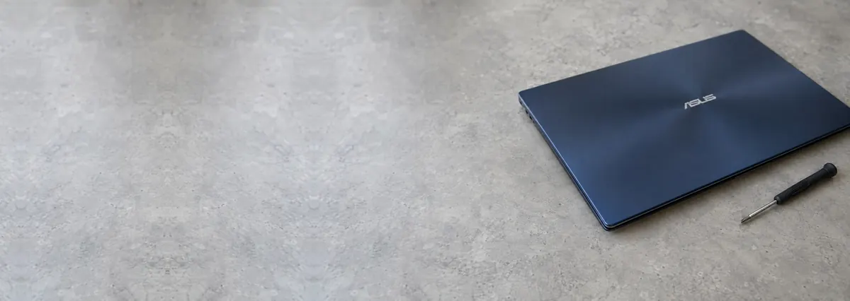 A closed dark-blue ASUS laptop beside a screwdriver on a concrete-effect workbench