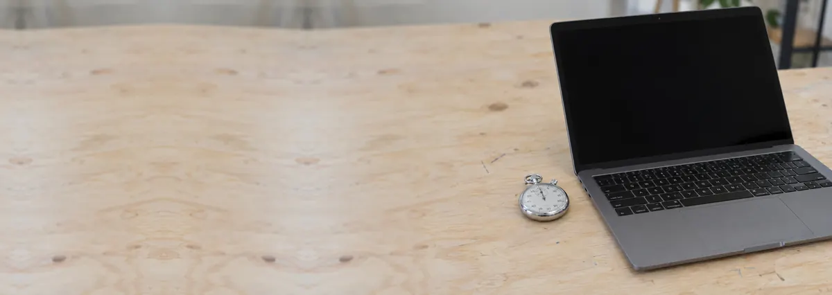 An open grey laptop with its screen off beside a small stopwatch on a plywood bench