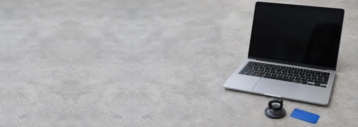 An open silver laptop with its screen off beside a suction handle and a pry card on a concrete-effect workbench