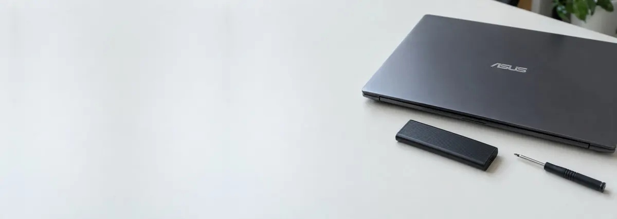 A closed ASUS laptop beside a slim black SSD enclosure and a screwdriver on a white bench