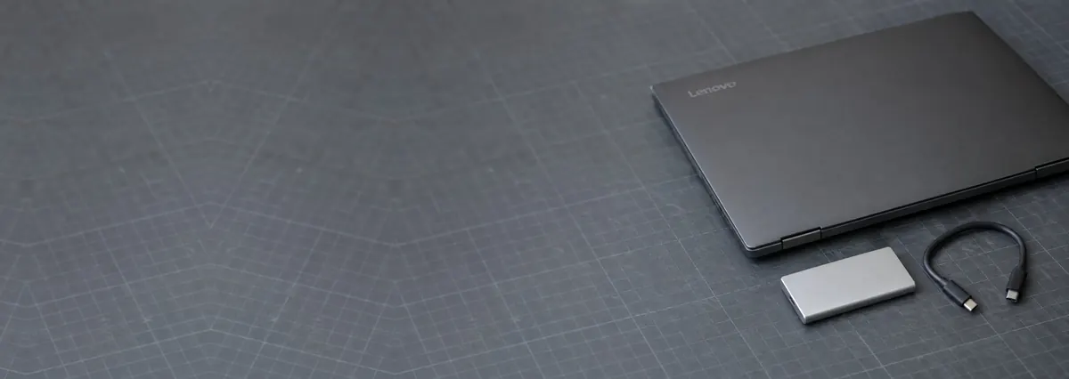 A closed Lenovo laptop beside a slim silver SSD and a USB-C cable on a slate-grey workbench