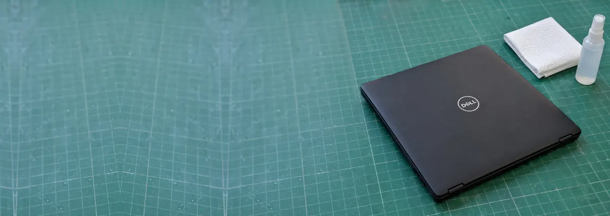 A closed black Dell laptop on a green cutting mat beside a paper towel and a spray bottle