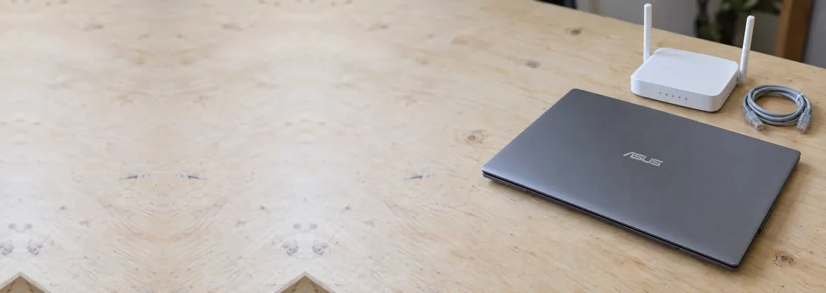A closed ASUS laptop beside a small white router and a network cable on a plywood bench