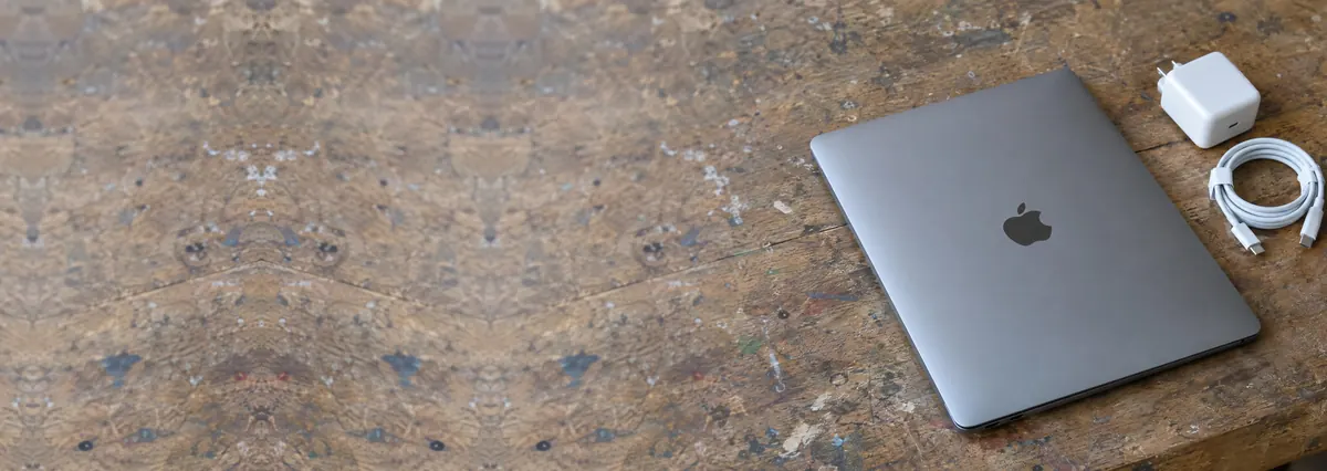 A closed space-grey aluminium Apple MacBook with its white charger and cable on a wooden bench