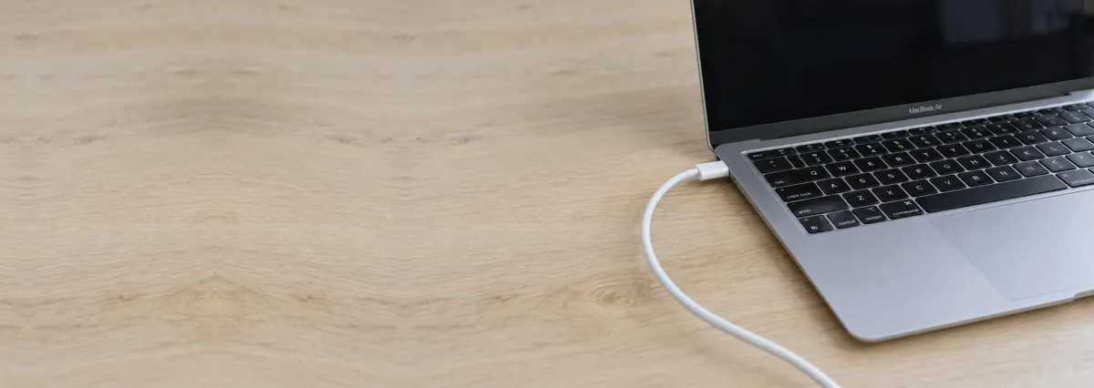 An open silver Apple MacBook with a white charger cable plugged in on an oak desk top