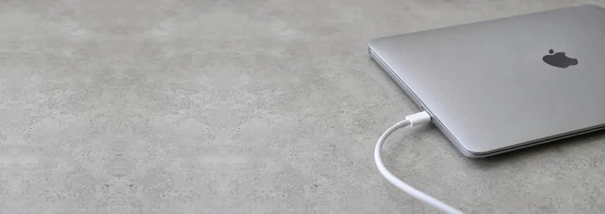 A closed silver Apple MacBook with a white USB-C cable plugged into its side port on a concrete-effect workbench