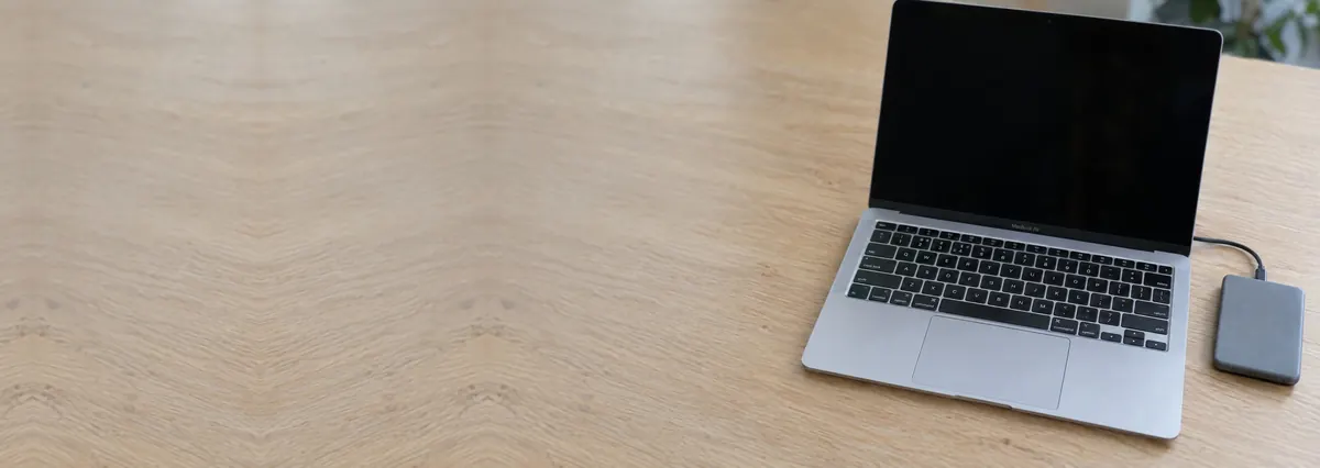 An open silver Apple MacBook with its screen off beside a small external drive on an oak desk top
