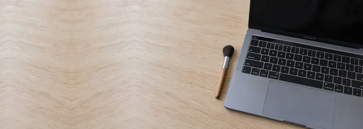 An open Apple MacBook with its keyboard in focus beside a brush on an oak desk top