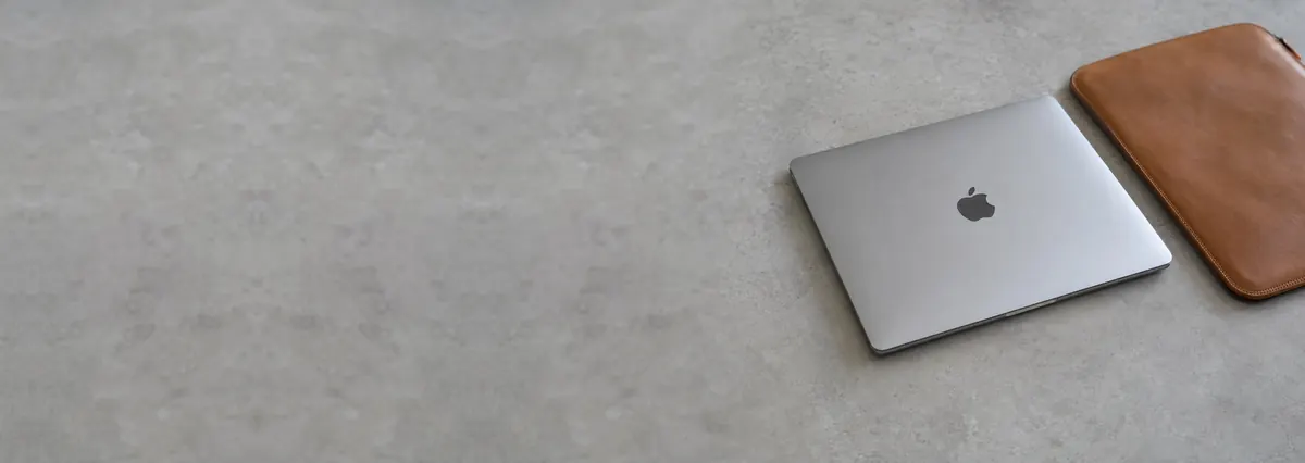 A closed silver Apple MacBook beside a leather sleeve on a concrete-effect workbench