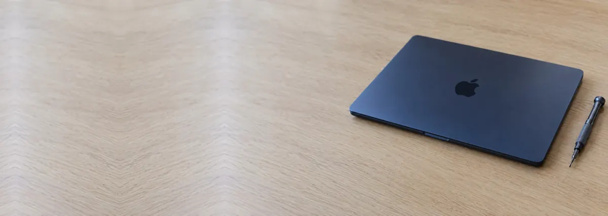 A closed midnight-blue aluminium Apple MacBook beside a screwdriver on an oak desk top