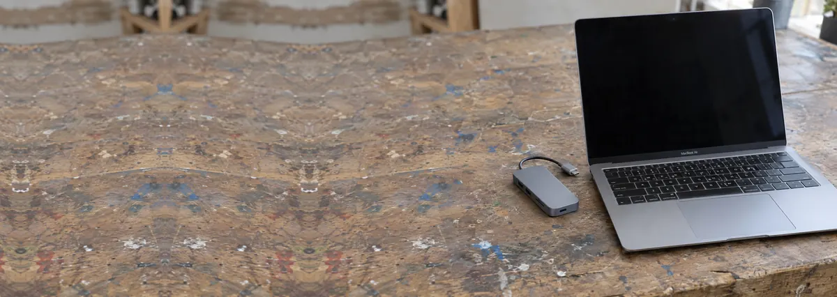 An open silver Apple MacBook with its screen off beside a small USB-C hub on a wooden bench