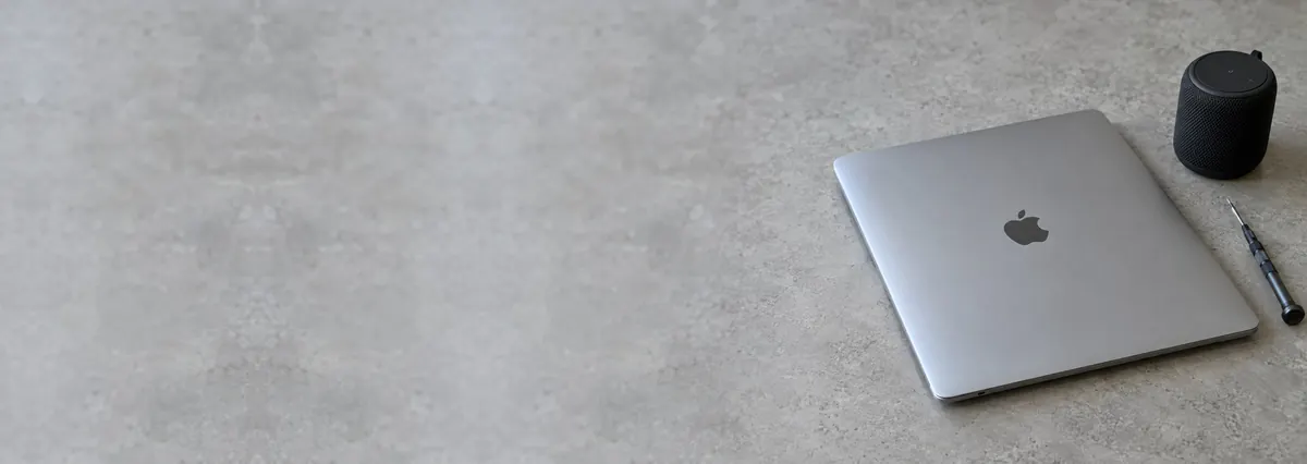 A closed silver Apple MacBook beside a small black speaker and a screwdriver on a concrete-effect workbench