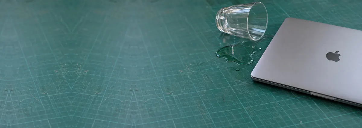 A closed Apple MacBook beside a small spill of water and a tipped glass on a green cutting mat