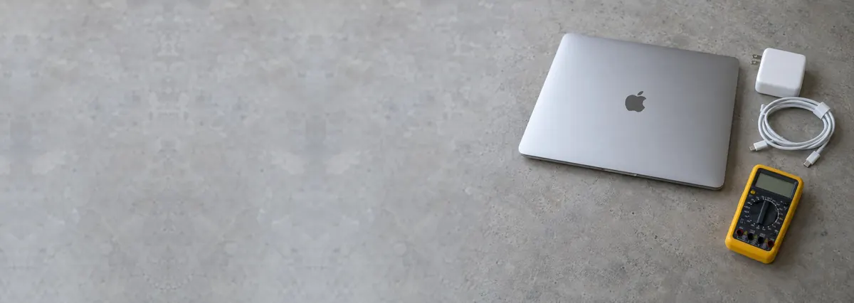 A closed Apple MacBook beside an unplugged charger and a multimeter on a concrete-effect workbench