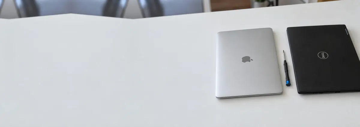 A closed Apple MacBook and a closed Dell laptop with a screwdriver between them on a white bench