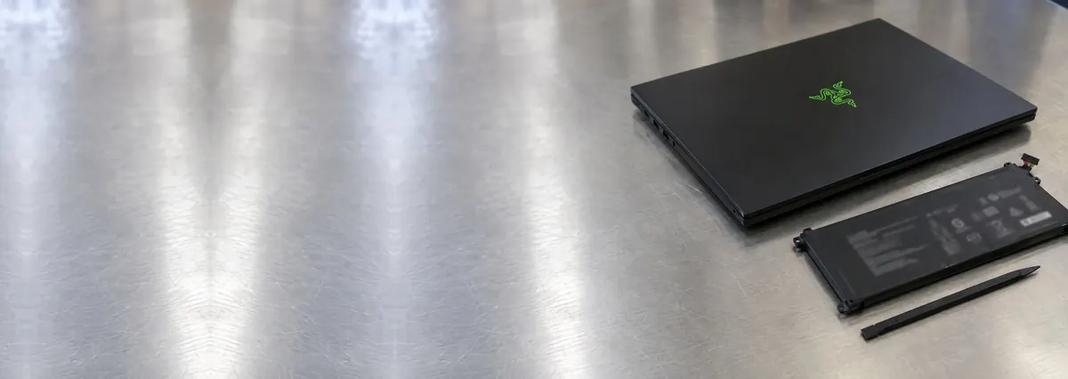 A closed black slim Razer Blade gaming laptop beside a flat battery pack and a spudger on a steel workbench