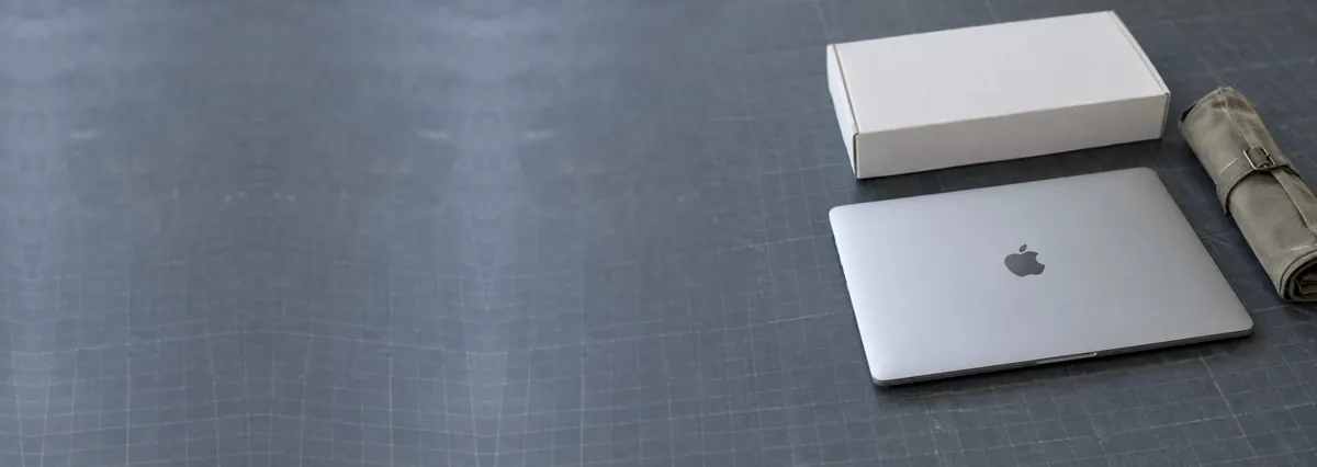 A closed Apple MacBook beside a plain white box and a toolkit roll on a slate-grey workbench