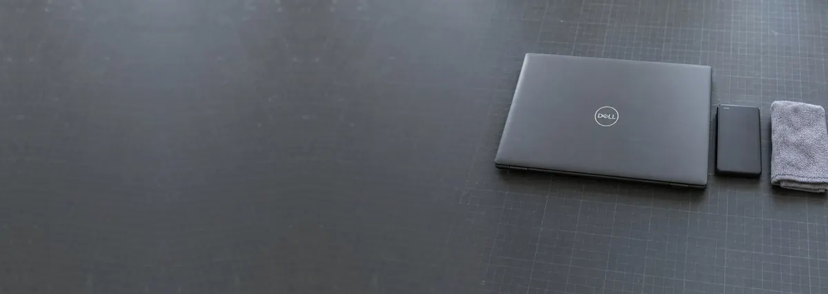 A closed Dell laptop and an external hard drive beside a folded towel on a slate-grey workbench